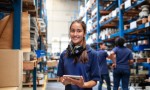 Portrait of a female factory employee with a digital tablet looking at camera and smiling. Woman warehouse worker with people in background.