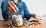 The woman hand is putting a coin in a glass  bottle and a pile of coins on a brown wooden table,Investment business, retirement, finance and saving money for future concept.